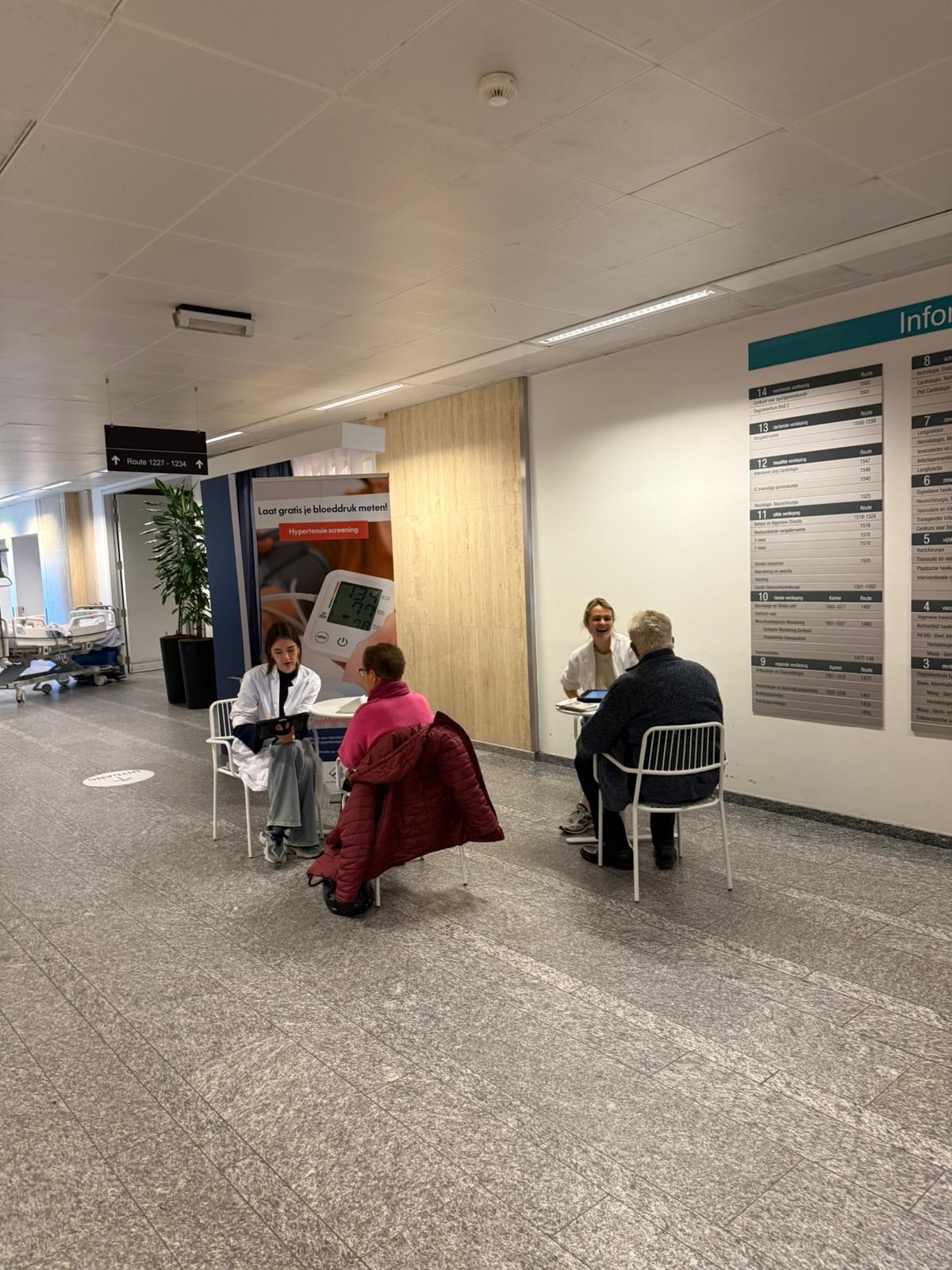 Deux ambassadrices prennent la tension de patients dans un couloir d'hôpital, roll-up « Laat gratis je bloeddruk meten! »